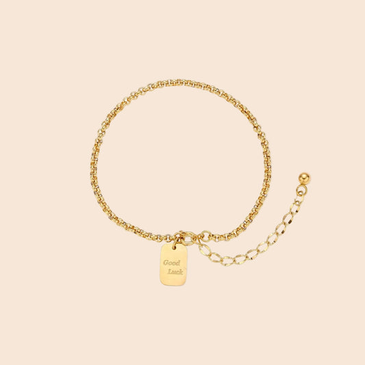 Delicate 18K gold-plated chain bracelet with Good Luck engraved rectangular pendant on cream background