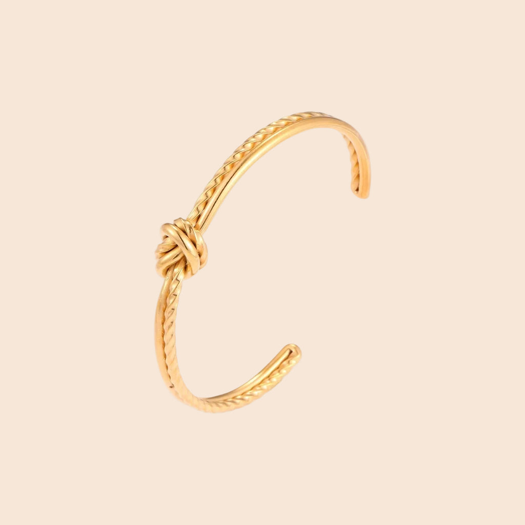 Gold cuff bracelet with twisted rope texture and decorative knot centerpiece on cream background.