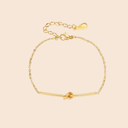 Delicate gold chain bracelet with minimalist bar and centered knot detail on cream background