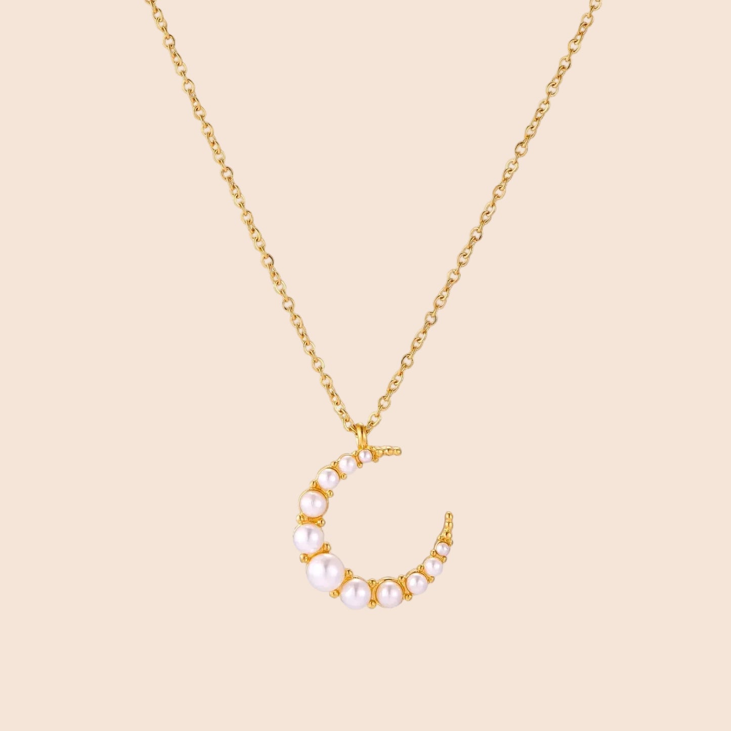Gold crescent moon pendant with graduated white pearls on delicate chain against cream background