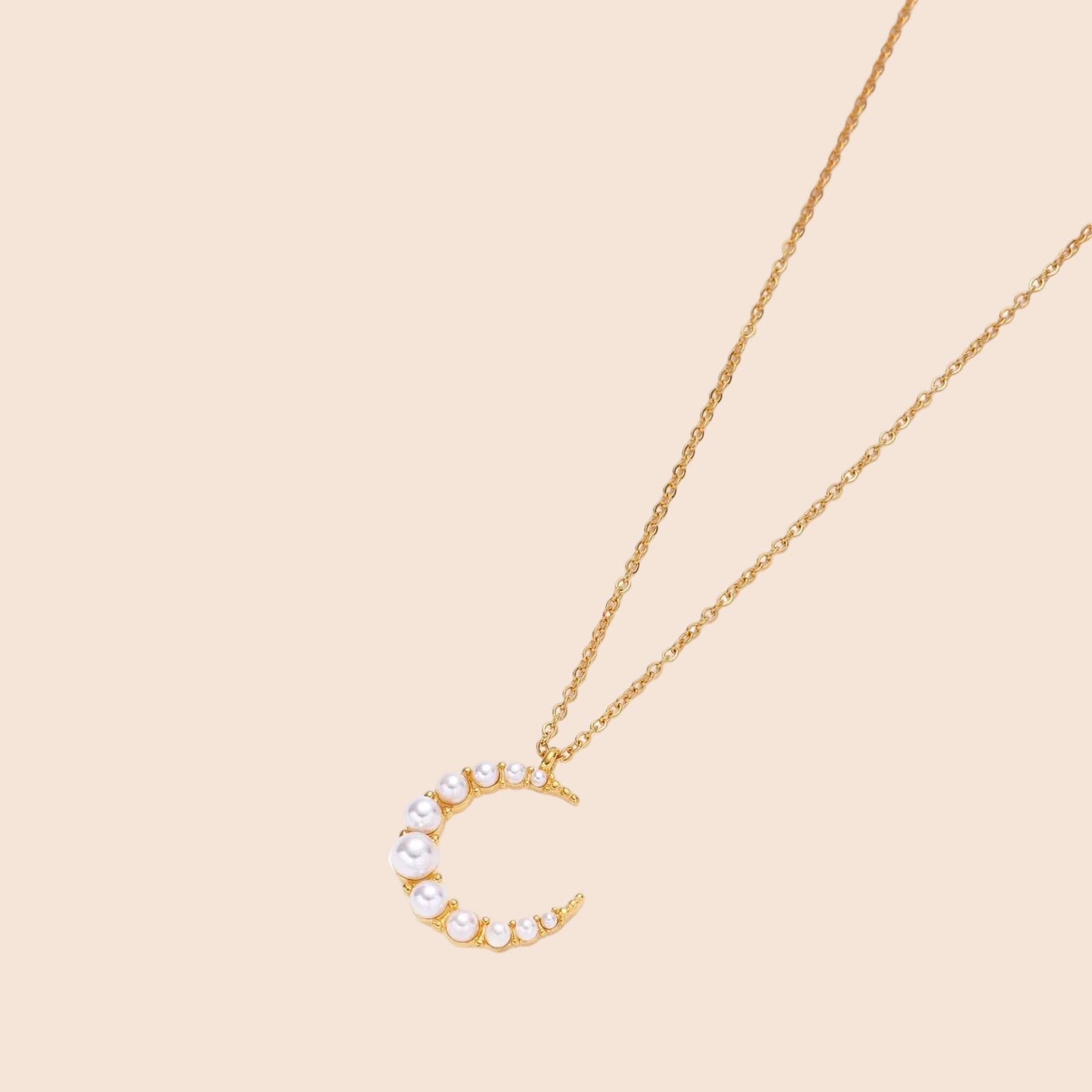 Gold crescent moon pendant with graduated white pearls on delicate chain against cream background