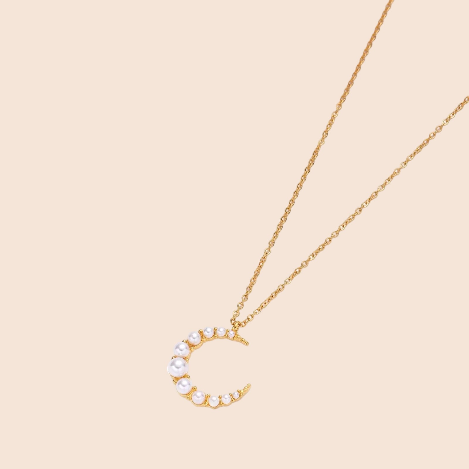 Gold crescent moon pendant with graduated white pearls on delicate chain against cream background