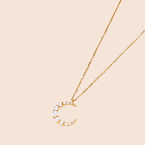 Gold crescent moon pendant with graduated white pearls on delicate chain against cream background