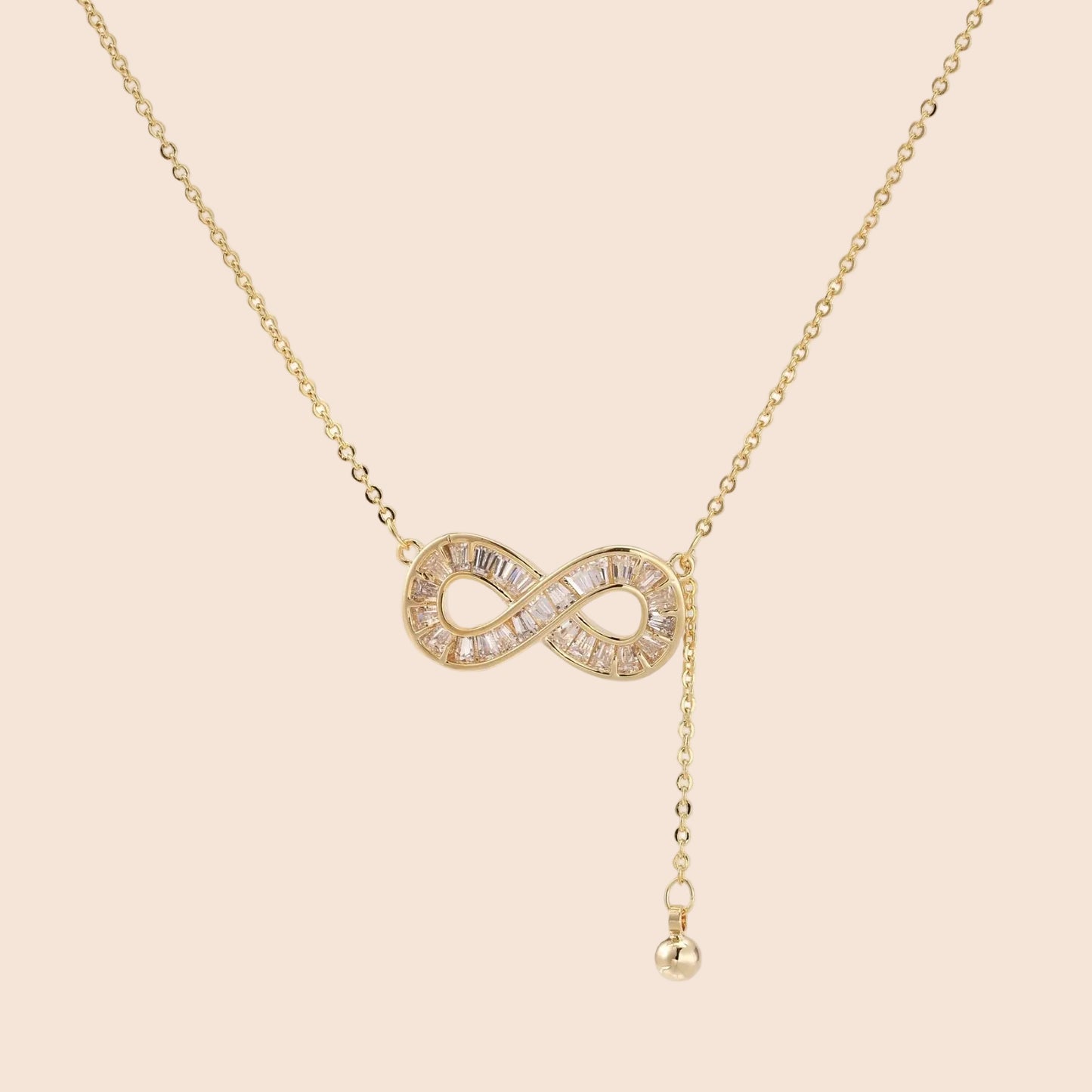 Gold infinity pendant with baguette crystals on adjustable chain against cream background.