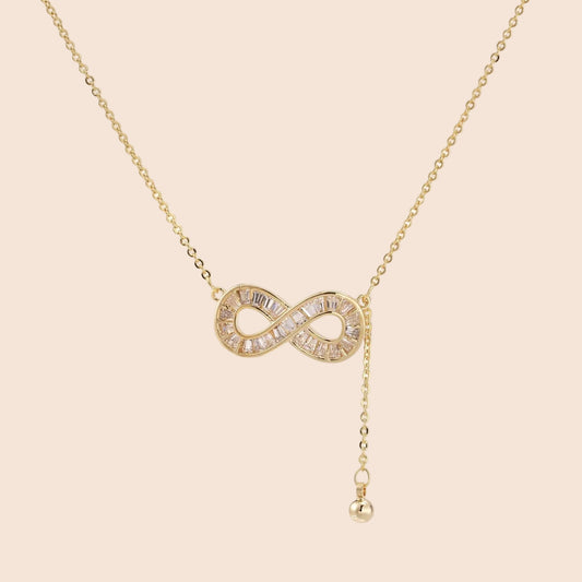 Gold infinity pendant with baguette crystals on adjustable chain against cream background.